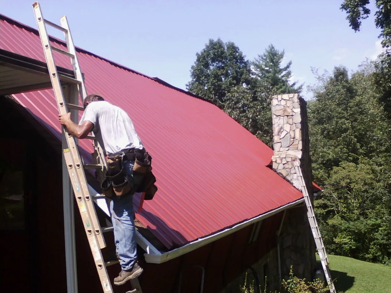 Roofer on a ladder beside a red metal roof and stone chimney — Soffit & Fascia Repair by Roofing Brothers in Dennis