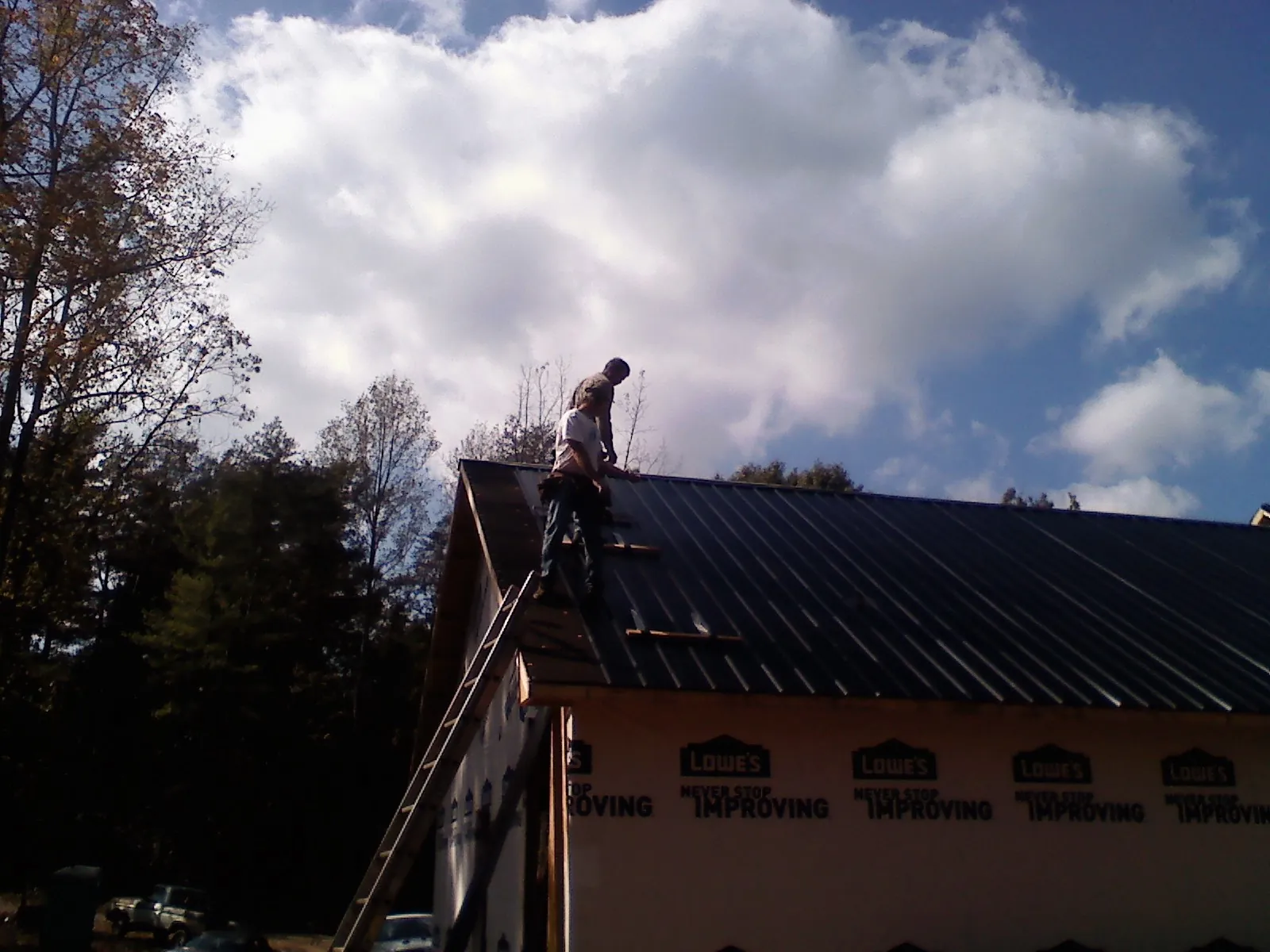 Roofer fastening dark metal panels high against a cloudy sky, from a Roof Inspection job in Dennis